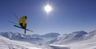 Skier in a yellow jacket jumping with crossed skis against sun and snowy peaks, Ski Classics offer near Hotel Bellerive Zermatt.
