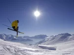 Skier in a yellow jacket jumping with crossed skis against sun and snowy peaks, Ski Classics offer near Hotel Bellerive Zermatt.