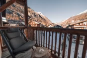 Wooden balcony of the Cosy Chic Room with a lounge chair and stump table, looking over Zermatt and snowy mountains.