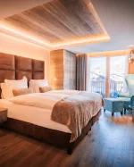 Chambre Premium Chic avec lit double, fauteuil turquoise, plafond en bois et vue balcon sur chalets et montagnes, Hotel Bellerive Zermatt.