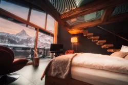 Sky Room am Abend: Bettkante und roter Sessel vor der Glasfront, Holztreppe mit Licht und Matterhorn im Abendrot, Hotel Bellerive Zermatt.