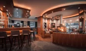 Hotel bar with a timber counter, grey stools and a round glass fireplace in the lounge at Hotel Bellerive Zermatt.