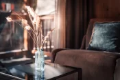 Room detail: ribbed glass vase with dried grasses on a coffee table, armchair and balcony in the background.
