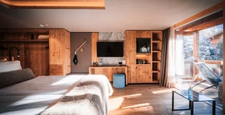 Chambre Deluxe claire, lambris bois, lit, mur TV et vue balcon sur neige et chalets – Hotel Bellerive Zermatt.