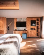 Helles Deluxe-Zimmer mit Holzwänden, Bett, TV-Wand und Balkonblick auf Schnee und Chalets im Hotel Bellerive Zermatt.