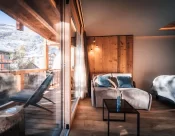 Bright sofa corner with glass doors to a wooden balcony and village views in the Panorama Deluxe Room at Hotel Bellerive Zermatt.