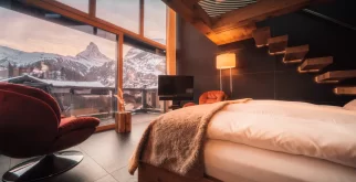 Sky Room mit weissem Bett, roten Sesseln und beleuchteter Holztreppe; durch die Glasfront Matterhorn im Abendlicht, Hotel Bellerive Zermatt.