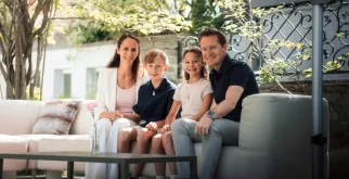 Smiling host family on a light outdoor sofa on the garden terrace with stone wall and trees, Hotel Bellerive Zermatt.