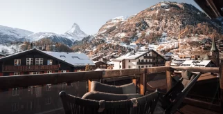 Balcon en bois avec deux chaises longues et rambarde vitrée, vue sur Zermatt enneigé et le Cervin – Hotel Bellerive Zermatt.