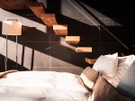 Schwebende Holzstufen an dunkler Steinwand über weissem Bett, Sonnenlicht und Schatten im Sky Room, Hotel Bellerive Zermatt.